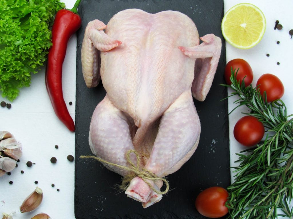 close-up-of-a-raw-whole-chicken-10842246 Overhead shot of a raw whole chicken surrounded by fresh vegetables and spices on a slate board.